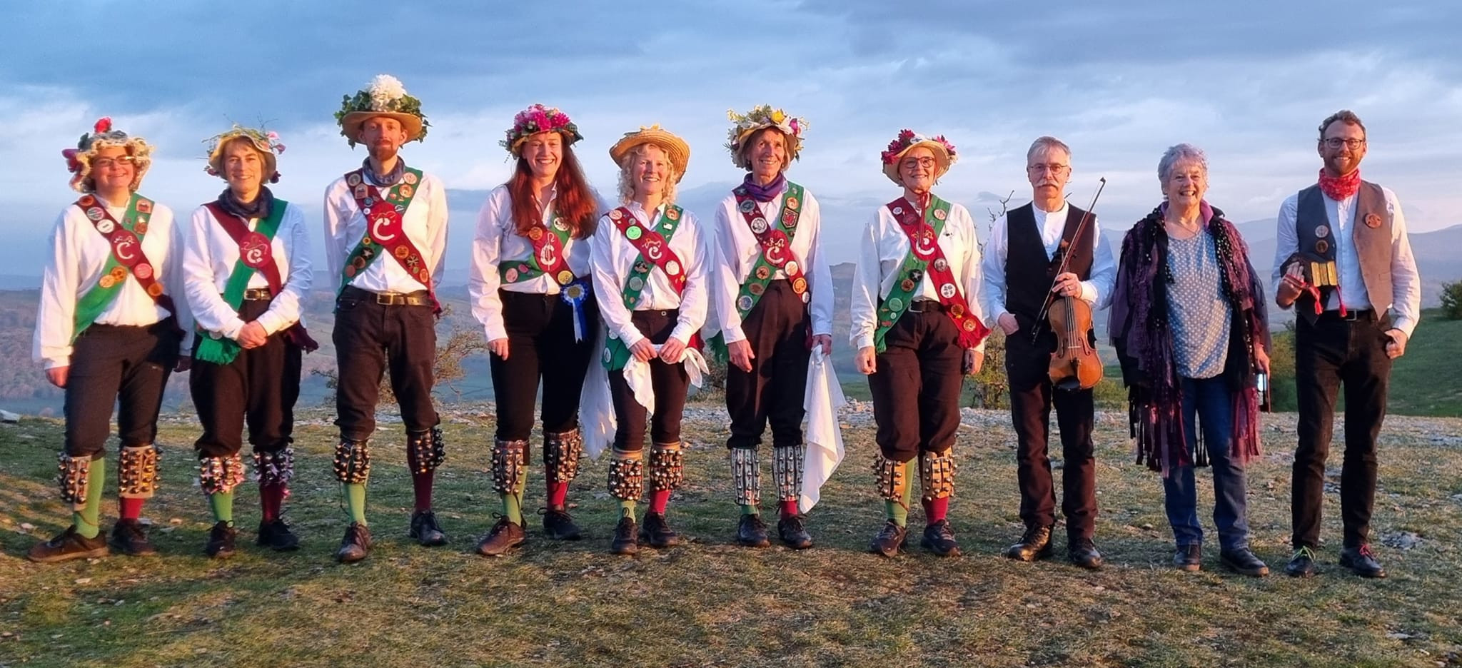 Seven jolly dancers and three musicians standing in a line on top of a Cumbrian fell soon after sunrise. They wear white shirts, black trousers, maroon and green baldricks and socks, and straw hats decorated with flowers.